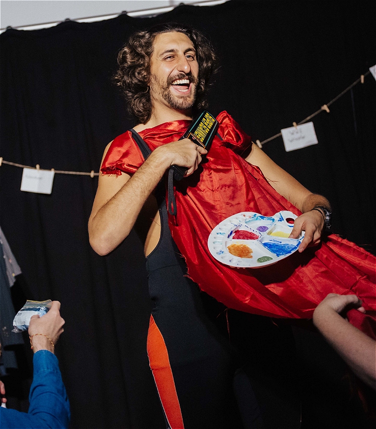 The performer is wearing a red fabric as a bib. He is holding a metal detector. An audience member is holding the other end of the fabric as a painters pallet is placed on the fabric.