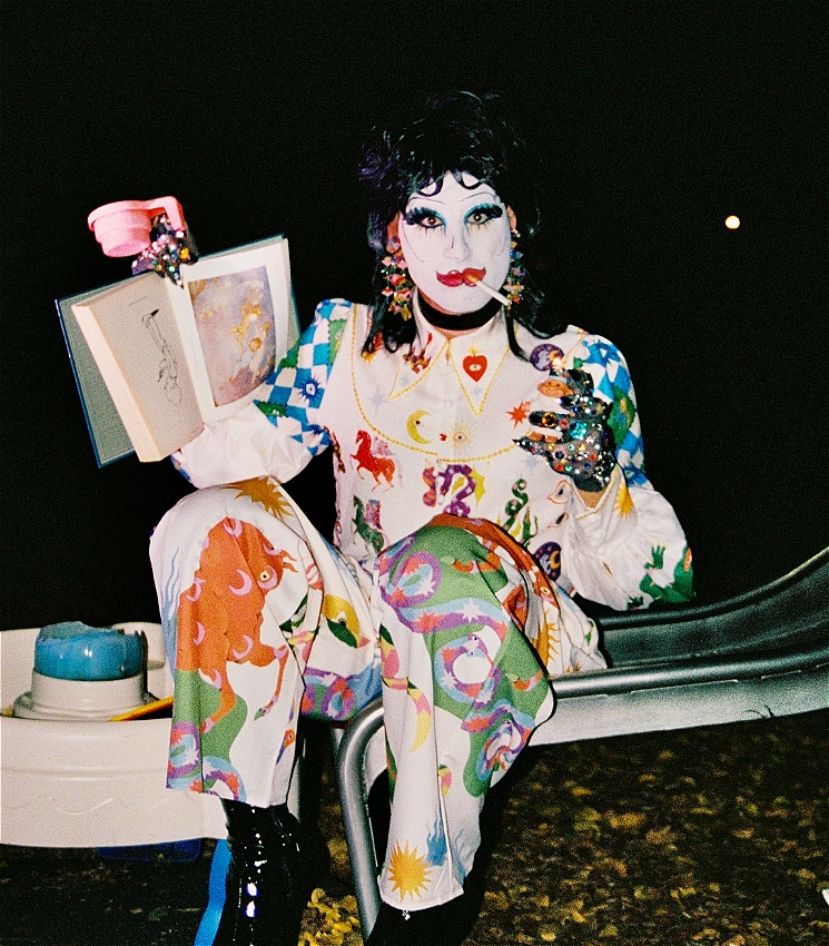 A drag artist in white clown make up is sitting at the bottom of a slide in a playground at night with a cigarette hanging out of their mouth. They are holding an open picture book and a plastic cup in one hand, and a lighter in the other hand. There are a few toys on the ground around them.