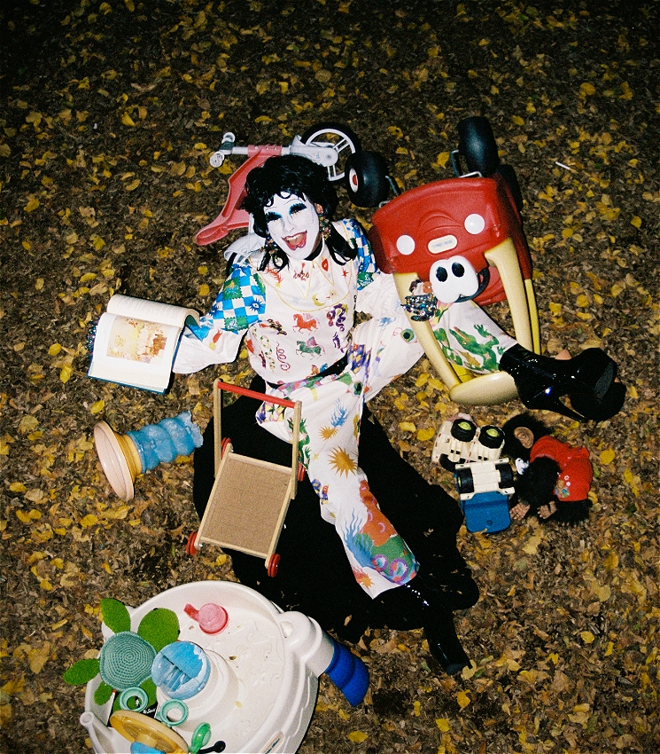 A drag artist in white clown make up and a manic smile is lying on the ground of an outdoor playground at night. They are surrounded by toys, including a book, small push-along bike and car, a plush monkey and turtle and some outdoor water toys. One of their legs is tangled through the openings of the push-along car toy.