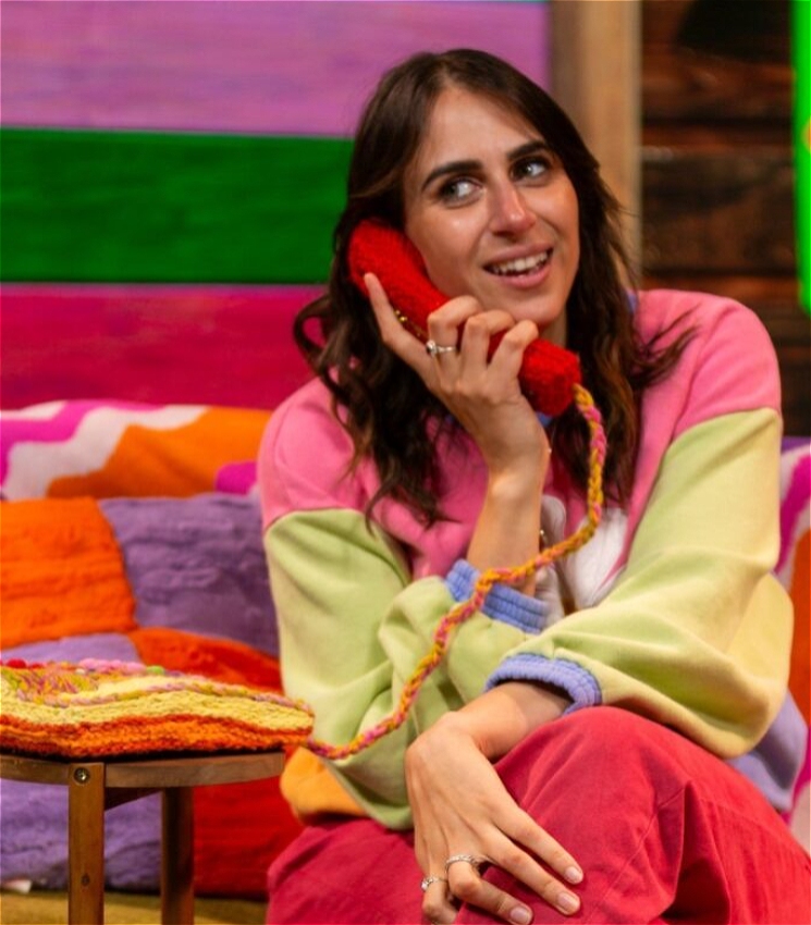 Ashley (a Caucasian woman with olive undertones who has brown wavy shoulder length hair and hazel eyes) sits on on a colourfully decorated couch. Behind her is an LED screen showing the interior of a wood-clad clubhouse with pink and green paint covering some panels. Ashley wears pink jeans, and a half pink, half light green jumper that has light purple cuffs at the sleeves and a big embroidered daisy in the middle of her chest. She holds a crocheted landline phone to her ear, looks to the side of the frame and wears a cheeky smile.