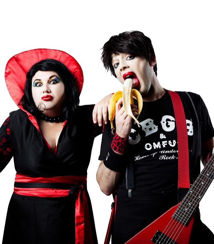 On the left is a woman with black hair and vibrant red-and-blue makeup. She wears black dress with a cherry-red sash around the waist, and a matching ruff that extends above her head. She wears a string of large black beads around her neck. On the right is a man with black hair and bright red lipstick, who is wearing a band t-shirt. He has a red electric guitar in one hand, and a ripe banana in the other, which he is about to eat. They are both rockstars.