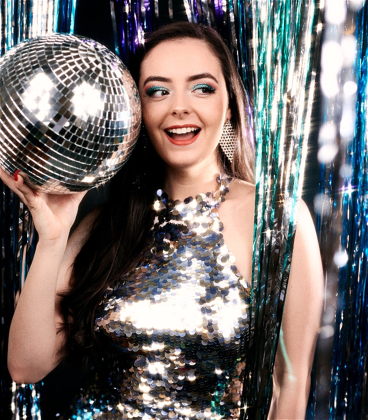 The artist wears a silver sequin sparkly dress. She is standing holding a disco ball in front of shiny streamers, looking towards the disco ball.