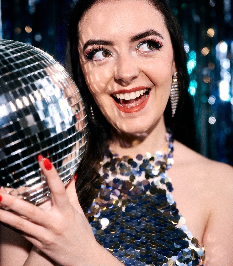 The performer holds a disco ball close to her face and looks away into the top right corner towards the light. She wears a sequin dress and is in front of a silver streamer backdrop.