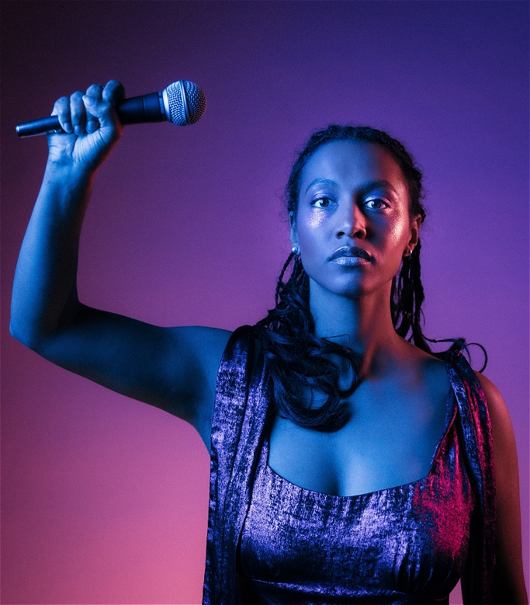 Person holds a microphone with one hand raised against a colorful gradient background, wearing a sleeveless top.