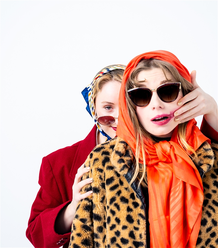 One blonde woman with smeared lipstick, sunglasses, a scarf and a fur coat looks shocked, while another blonde woman also in sunglasses and a scar, and a long red coat stands behind her menacingly. The woman in the red coat has her her left hand wrapped around the woman in front of her, and her right hand smearing the woman in front of hers lipstick in a way that suggests she had her hand over her mouth.