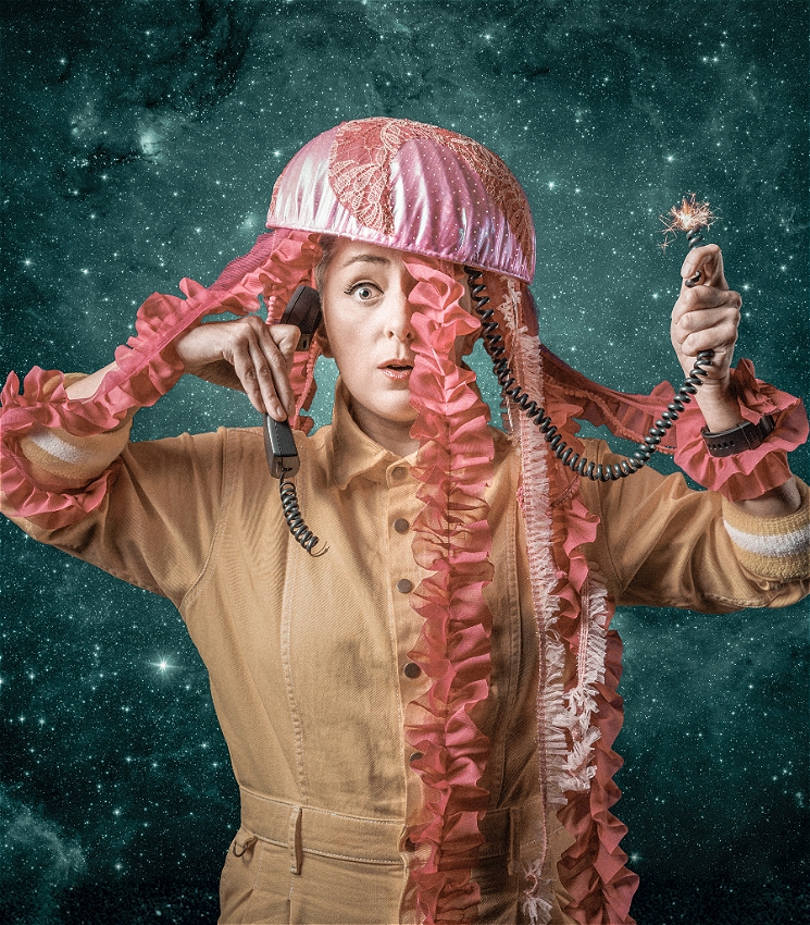 A person in a beige boilersuit wearing a jellyfish costume on their head and holding a telephone to their ear in front of a backdrop of stars.