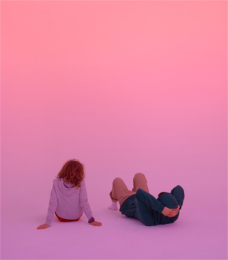 Against a bright pink background, two people sit on the floor with their back to the camera. One lies on their back, knees bent, with their hands holding their head up; the second sitting up straight. Each person's head is slightly tilted toward each other, but their faces are not seen.
