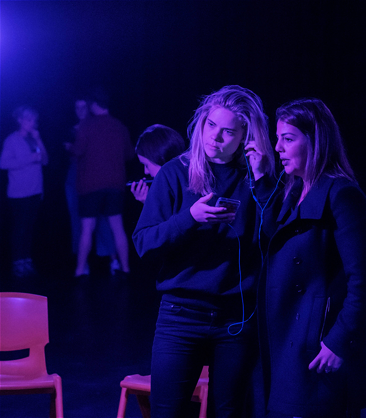 In the foreground, two people stand close together under dim theatre lights. They are listening to something on a phone through a shared set of headphones. In the mid-ground, a person is speaking into her phone. In the background, a group of three people stand together in a discussion.