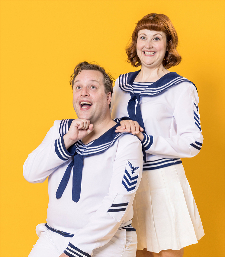 A man with short blonde hair wearing a blue and white sailor's outfit sits as a woman wearing an identical sailor suit stands behind him and rests her hands on his shoulder. Both have ridiculous smiles and they are against a yellow background.