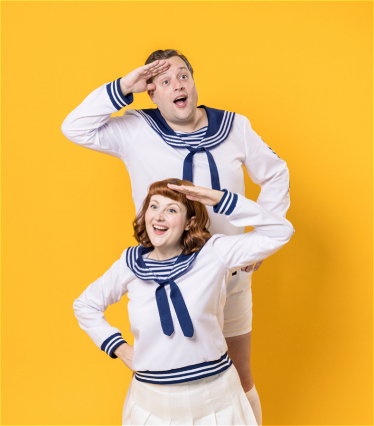 Two people, a man and a woman, are dressed in matching sailor-themed outfits with white tops and blue collars. They are both smiling and posing in a playful manner, with one hand raised to their foreheads in a salute. The background is a bright yellow color, adding to the cheerful and lively atmosphere of the image.