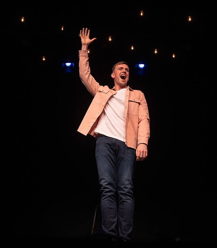 Tom, a white Irish man, stands on stage in a pink shirt. His hand is raised and his mouth is open.