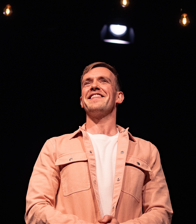 A close up of Tom, a white Irish man, stands on stage in a pink shirt smiling.