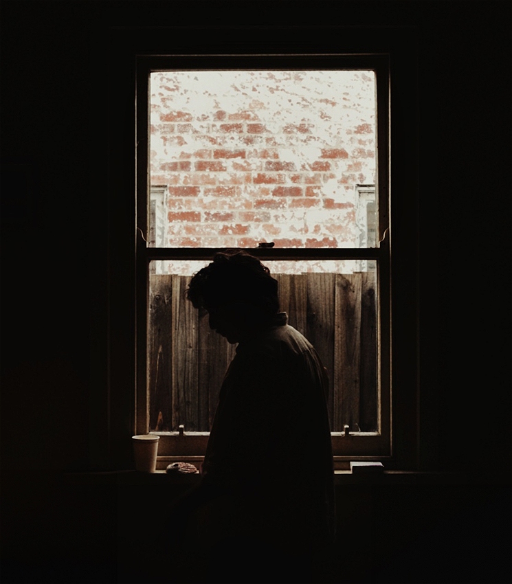 A completely dark room with a window in the center. The view is suburban - framing a wet wooden fence with a wall beyond, weathered white paint flaking, revealing the orangey red bricks beneath. A figure stands in silhouette, and we can just make out his medium length dark wavy hair and the texture on the back of the corduroy shirt he wears. On the windowsill, in the left corner, is an empty white paper cup, next to a palm-sized nautilus-like seashell.