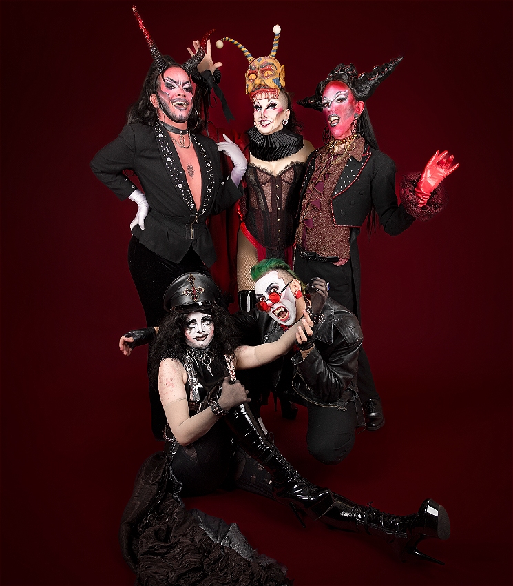 Five drag artists dressed in eclectic red and black clothing resembling demons in front of a blood red background. Three are standing behind two on the ground at the front, with one pretending to bite the arm of the other.