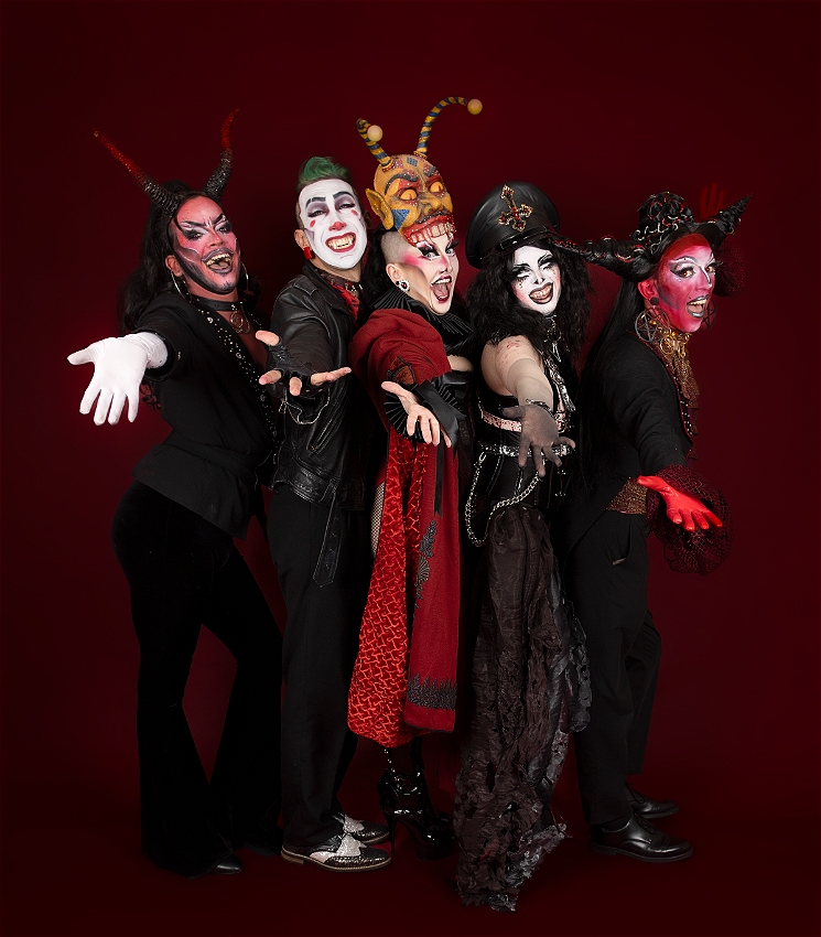 Five drag artists dressed in eclectic red and black clothing resembling demons in front of a blood red background. They are each standing back to back with their outer arms outstretched towards the audience resembling a cheesy 