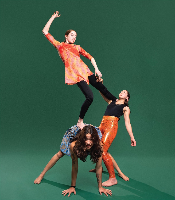 Three females dressed in black, orange, and blue are positioned in an unconventional pyramid in front of a bottle green backdrop. From bottom to top: the first female acts as the base of the pyramid. On her right, the second female leans backwards, counterbalanced by the third female with her left arm extended above her as she stares directly into the camera.