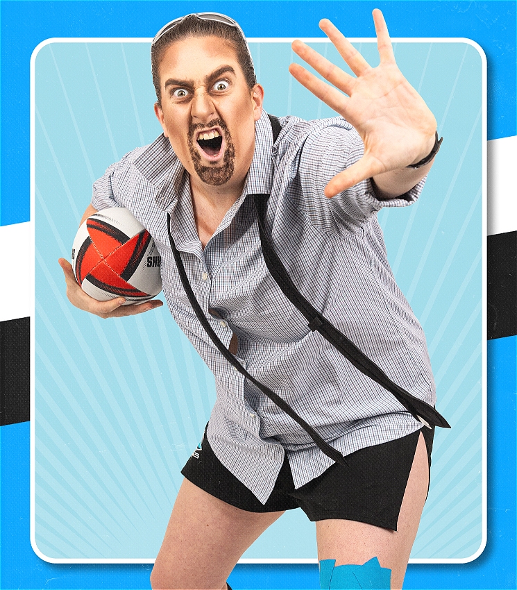 Drag King Scrum wears a button up shirt, undone tie with football shorts. He has an aggressive expression and is holding a football in one hand and the other hand out in front.