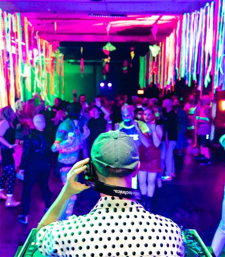 In the foreground is the back of a DJ he is playing on decks and holding his headphones, in front of him is a large colourful room with lots of people dancing.