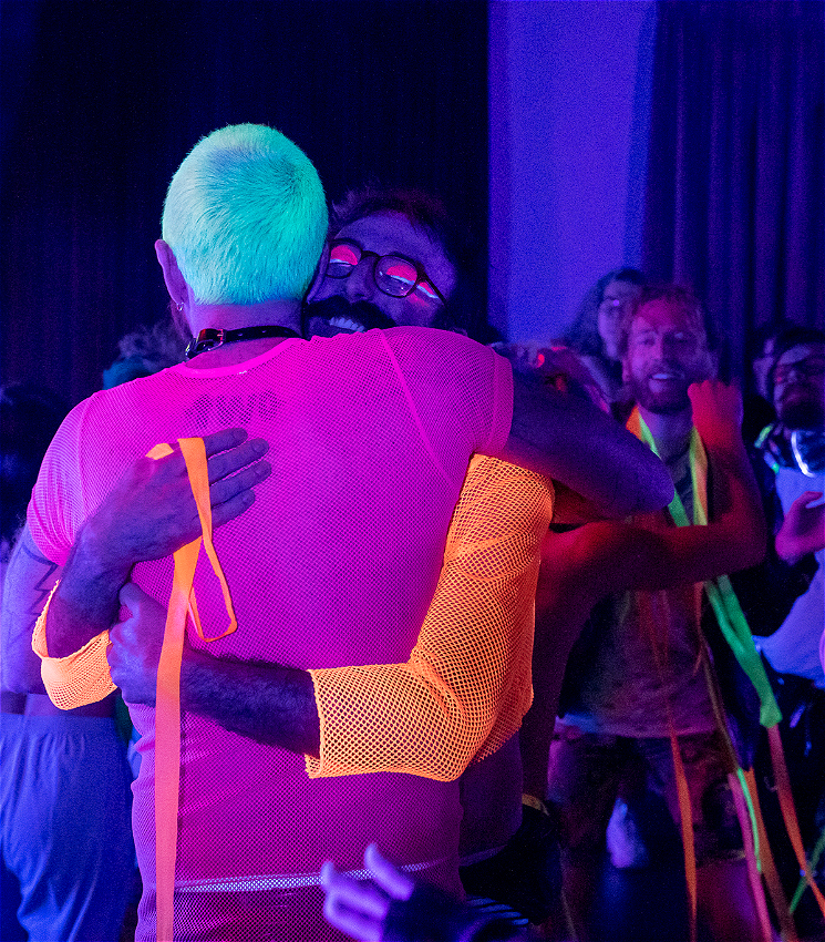 The room is bathed in florescent colours with many people dancing. The center of the image is two men embracing in a hug, the man facing away has bright green hair and a pink mesh top, the man facing forward has glasses and a orange mesh top.