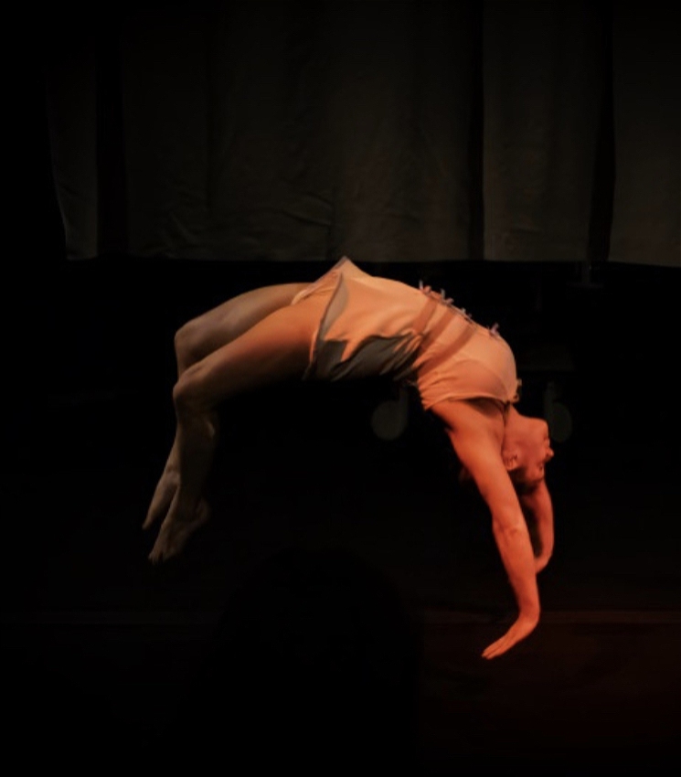 A close up shot of a performer doing a back handspring. She is in a backbend shape halfway through doing this skill. There is a redish tinge around the image and she is wearing a one piece costume that is inspired by a hospital gown/straight jacket.