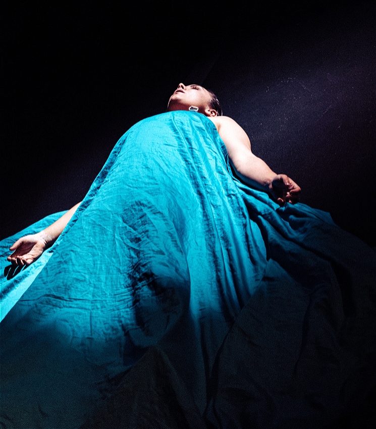 A person is lying on the ground with her arms out looking up. Her body is covered from her shoulders with a teal coloured sheet that is draping across her and the ground.