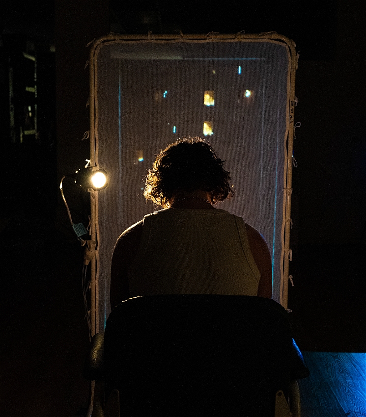 A man sits in a chair with his back facing the camera. He is wearing a white singlet. The space is dark, save for a single strong light emanating from his left side, illuminating his shock of wavy, messy hair, the silver chain on his neck glinting. In front of him is a window-sized piece of semi-transparent fabric, similar in texture to gauze, that is stretched on a white glossy metal frame. His head is bowed - the tone is somber and dramatic.