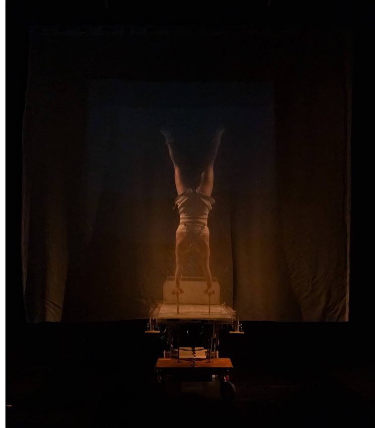 A performer is doing a handstand with her legs slightly apart. She is holding onto long canes that poke out of a hospital bed. She is behind a thin white projection screen and she is wearing white socks and a one piece costume.