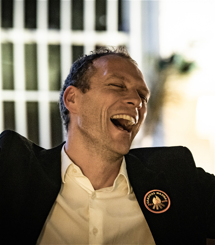 The image is close up of a person with short dark hair, singing joyfully. He is wearing a dark suit jacket over a white shirt. A round badge is pinned to his jacket, though the text on it is not fully legible. The background is softly blurred, featuring what appears to be vertical blinds and some indoor plants.