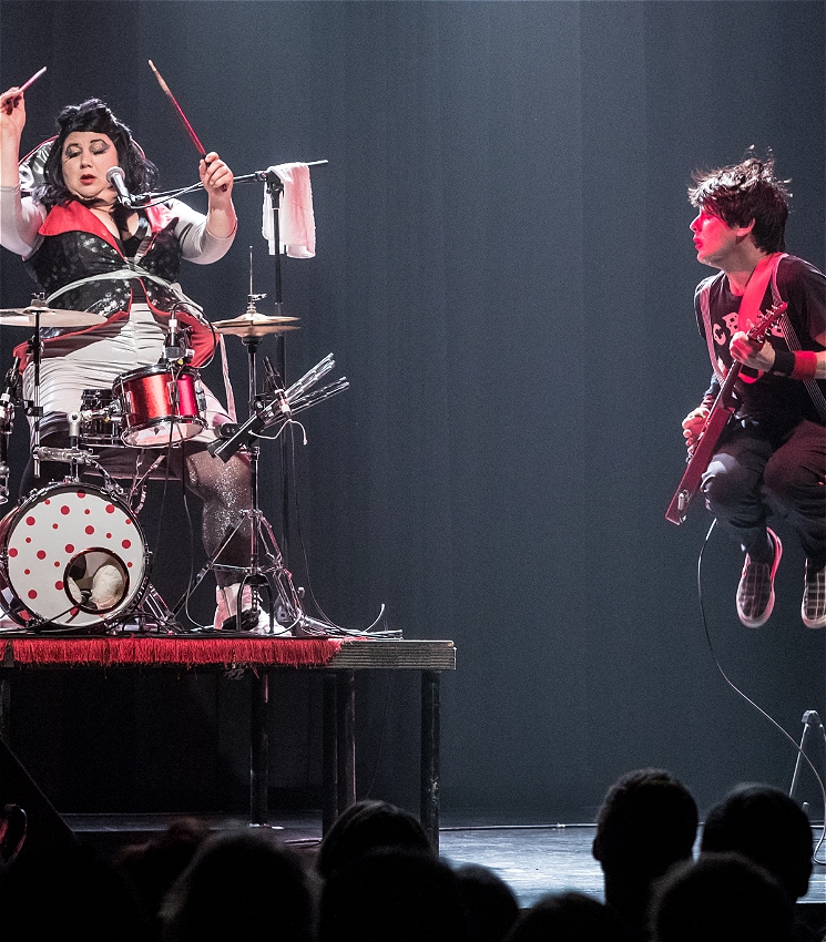 On the left is a woman with black hair and vibrant red-and-blue makeup. She is playing drums. On the right is a man with black hair and bright red lipstick, who is wearing a band t-shirt. He has a red electric guitar and is in a mid air leap.