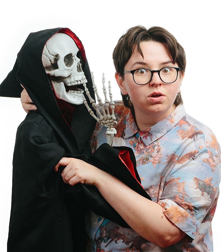 Comedian Maddy Weeks is holding a skeleton, draped in a black cloak, resembling the grim repaer. Maddy is a non-binary comedian with a mullet, brown hair and black glasses. They are wearing a button-up cherub-print shirt and brown pants. Maddy has a shocked look on their face and the skeleton is positioned to appear like it's whispering something to them.