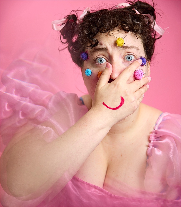 Person with blue eyes and brown curly hair in two buns with pink ribbons wearing a pink organza dress. They have colourful pom poms stuck to their face with a concerning expression in their eyes. Their hand covers their mouth with a red smile drawn on it, as they are framed in pink background that blends with their dress colour.