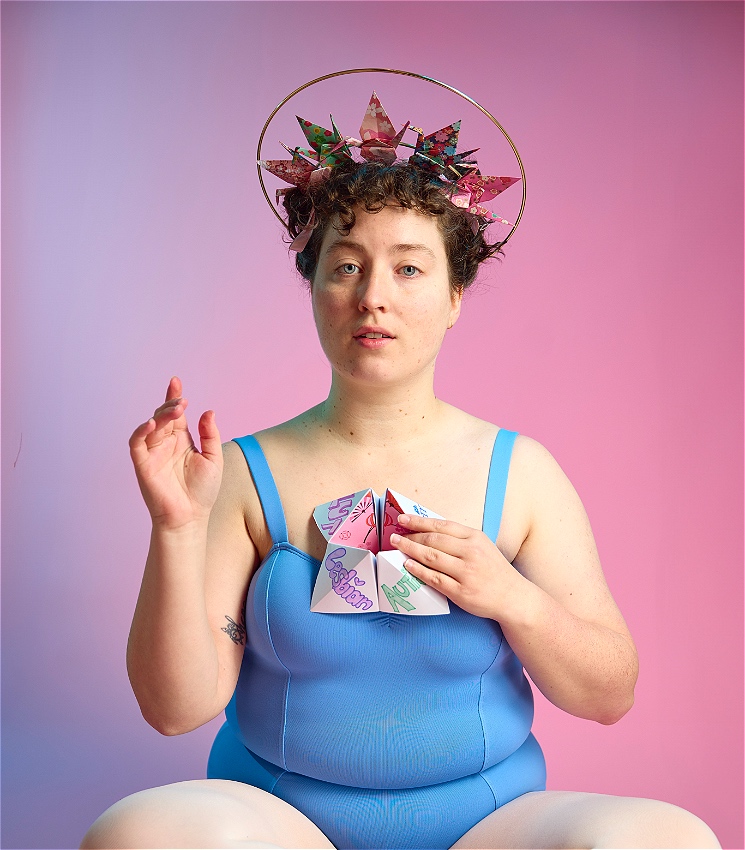 Person wearing a blue leotard, and a halo made from origami paper cranes, in a pose mimicking a statue of Mary holding an origami chatterbox with the words lesbian and autistic written on there.