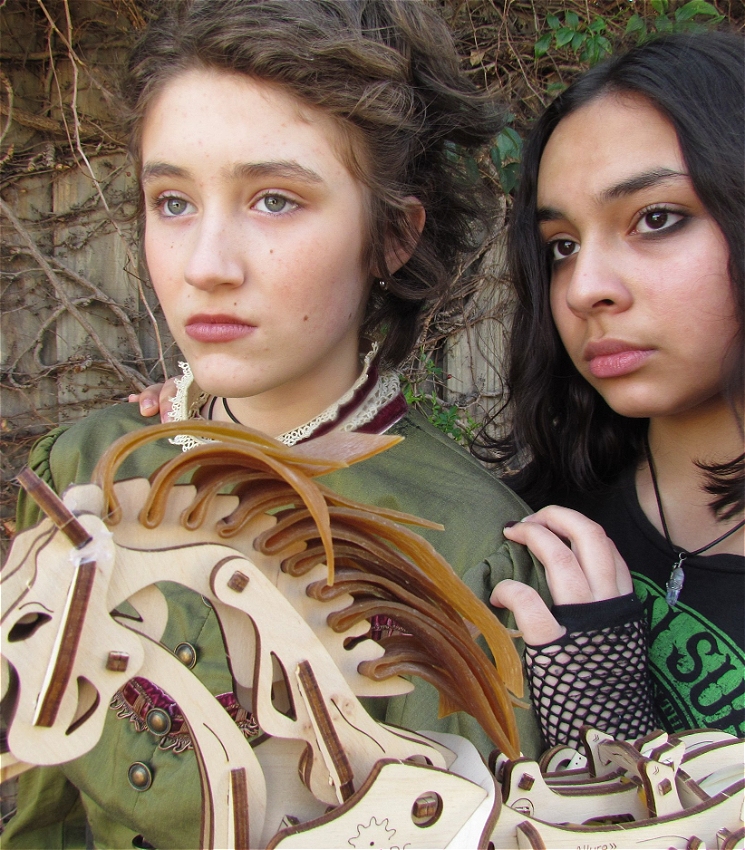 Two young women hold a wooden horse and look off into the distance. One is wearing Victorian clothing and one is dressed in modern clothing. They are touching, as friends.