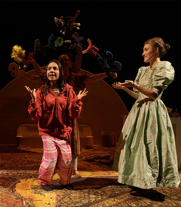 Two young women, one in modern pyjamas and one in Victorian dress stand together on a rug. There are puppets of numbers floating above them and they appear to be on stage. The ground is covered in Persian rugs.