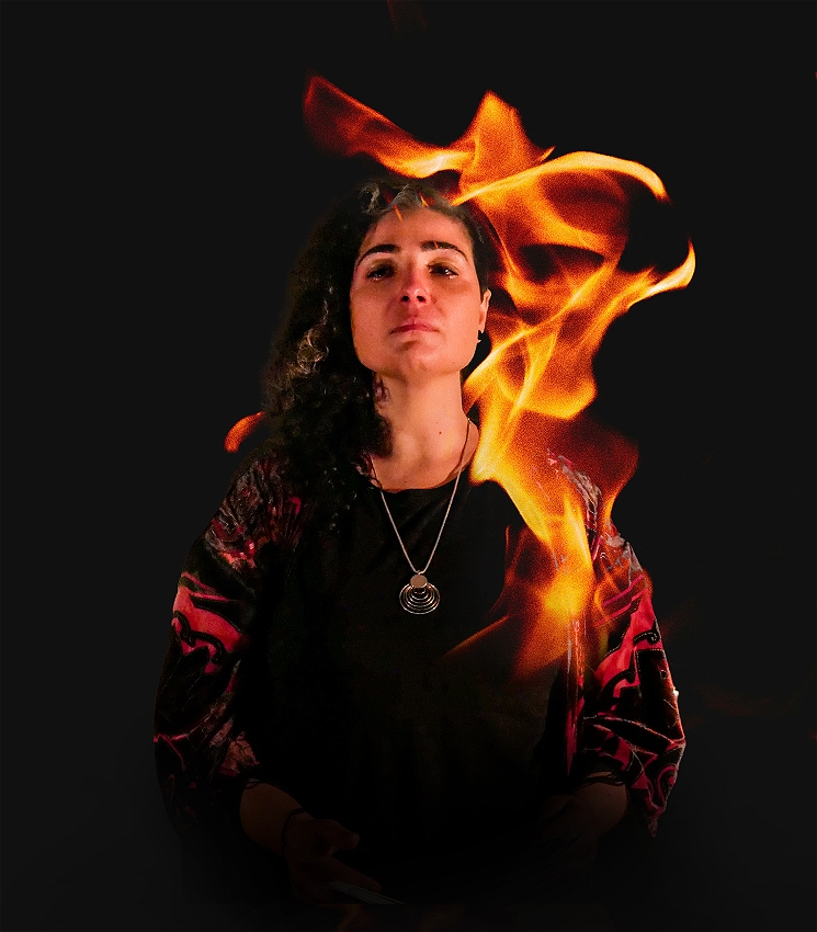 A powerful woman with long curly black hair stands fiercely gazing into the viewers eyes, with arms by her sides, and flames surrounding one side of her, framing her face.