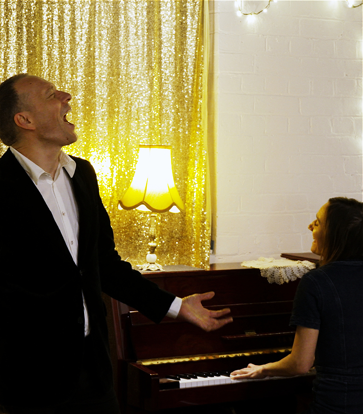 The image shows two people in a warmly lit room. To the left, the person is dressed in a dark suit and white shirt, and is singing passionately. To the right, a person is sitting at a piano, smiling, with their hands on the keys. In the background, there is a gold sequin curtain and a lamp with a yellow shade.