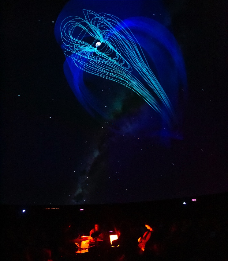 Musicians sit below a dark, starlit cosmos projected on the domed planetarium ceiling, their faces and instruments (cello, drums... ) aglow from the iPad screens they are reading from. Above them blue, aqua and green graphics emerge from the image of a moon, creating a giant jelly-fish like effect across the night sky.