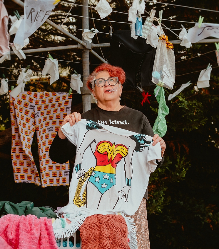A person wearing glasss stands under a hills hoist clothes line, holding a wonder woman T-Shirt. She has a clothes peg in her mouth.