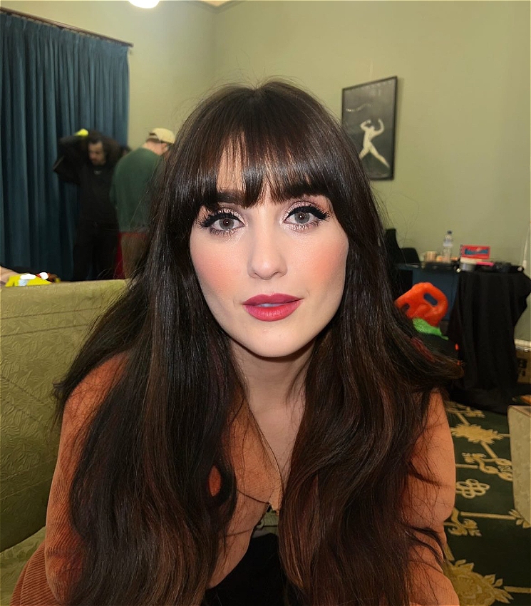 A woman with dark brown very long hair and a fringe looks into the camera. She has glamorous makeup on with bright pink lips and a brown cord jacket. She is sitting on a green couch and appears to be in a green room.