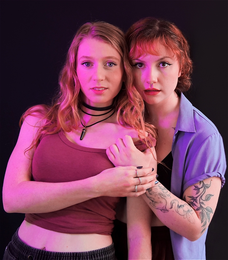 Two young people standing closely together against a plain black background with soft, coloured lighting that creates a blend of pink and purple tones on their skin and clothes. The person on the left is smiling slightly towards the camera, has long, wavy blonde hair and is wearing a mauve sleeveless crop top with a black choker necklace. The person on the right has short, wavy red hair and is wearing a lavender short-sleeve button-up shirt over a black bra. They have tattoos on their arms and are also looking directly at the camera with a serious expression. Their left arm is wrapped around the other person's shoulders, holding them close, while their right hand is slightly pulling the strap of the other person's shirt off their shoulder.