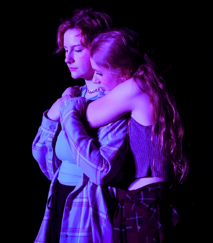 Two people standing close together against a black background, illuminated by purple lighting. One person, on the left, has short hair and is wearing a light blue top under an open, long-sleeved plaid shirt. Their gaze is on the floor and their hands are clasped together near their chest, over the other person’s arm. The other person, on the right, has long hair and is wearing a sleeveless, ribbed top. They are hugging the first person from behind, resting their head on the other person’s shoulder. The overall mood appears to be sombre, as if the hug is an apology.