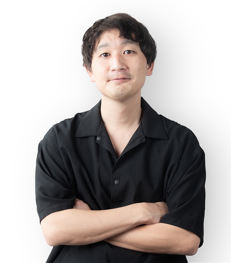 A Japanese man with dark hair smiles at the camera, folding his arms.
