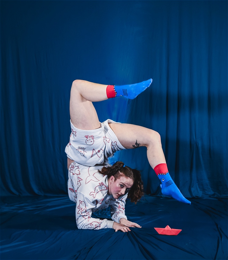 An acrobat in a contortion elbow stand, supporting their entire body weight in the air by their elbows, wears white clothing covered in hand drawn sea creatures at the centre of the image. Next to them is a small red paper boat on the floor. The back drop is blue.