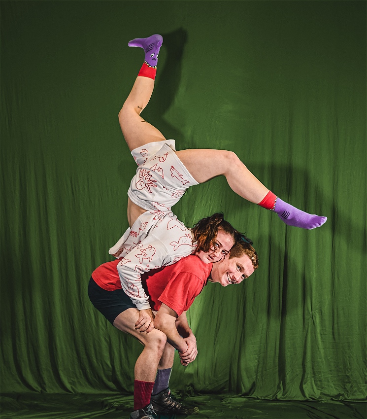 An acrobat dressed in red is leaning forward while an acrobat dressed in white performs a contortion chest stand on top of their back. The back drop is green, both appear excited.