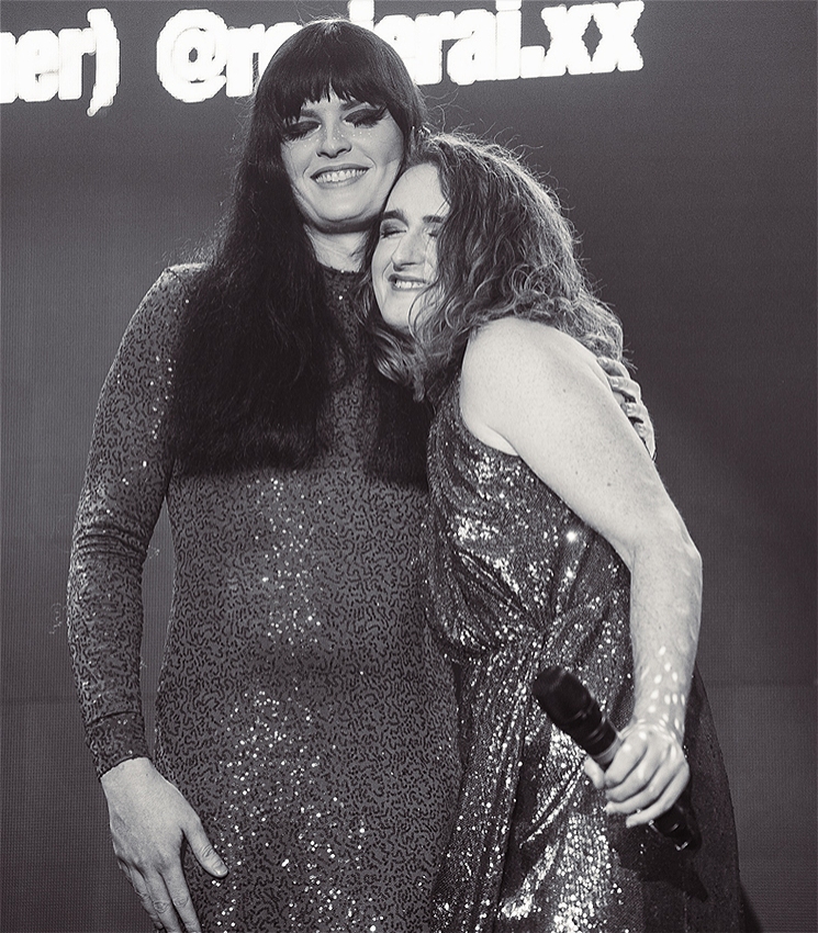 A photo of two women in black and white on stage wearing sequin gowns, smiling and hugging