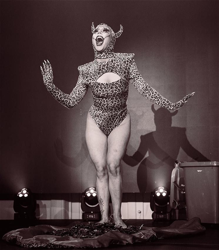 Drag and circus artist Themme Fatale pictured in black and white, they are wearing a leopard print body suit and devil horn headpiece, and their expression is a playful big smile with the clown-like makeup.