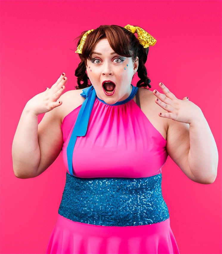 A white woman with brown hair in two pigtails stands in front of a pink backdrop. She wears a pink dress with a blue sparkly waist. Her hands are by her face and her mouth is open in an 'O'.