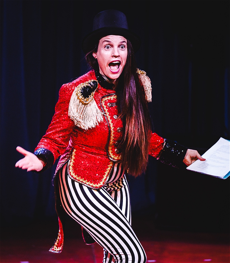 A white woman with brown hair wears a red glittery jacket in the style of a ringmaster and black and white striped pants. She holds a clipboard, hands extended out to her sides and has wide open eyes and mouth.
