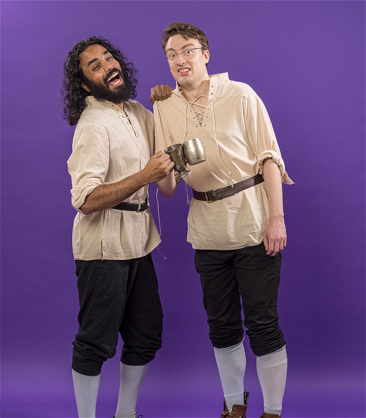 The image shows two people dressed in off-white shirts with their sleeves rolled up, black trousers, brown leather belts, white stockings and brown shoes in front of a purple background. They are holding steins in hand, struck together. One of them has a happy expression and the other an awkward.