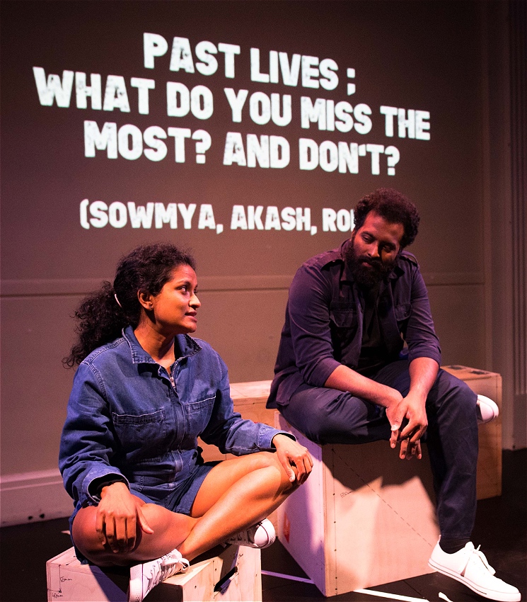 A man and woman sitting on wooden boxes talking to each other. The woman is sitting crossed legged and the man is sitting slightly above with one foot tucked underneath his other leg. Behind them text is projected against a back wall reading: 